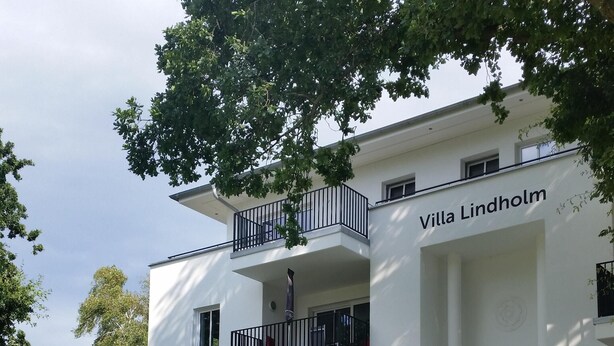 Un edificio moderno de dos pisos llamado "Villa Lindholm", con un balcón, rodeado de exuberante follaje y macizos de flores.