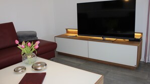 Un salón moderno con un televisor de pantalla plana sobre un mueble de madera, un sofá rojo, un florero de vidrio con flores rosadas y una mesa de centro blanca.