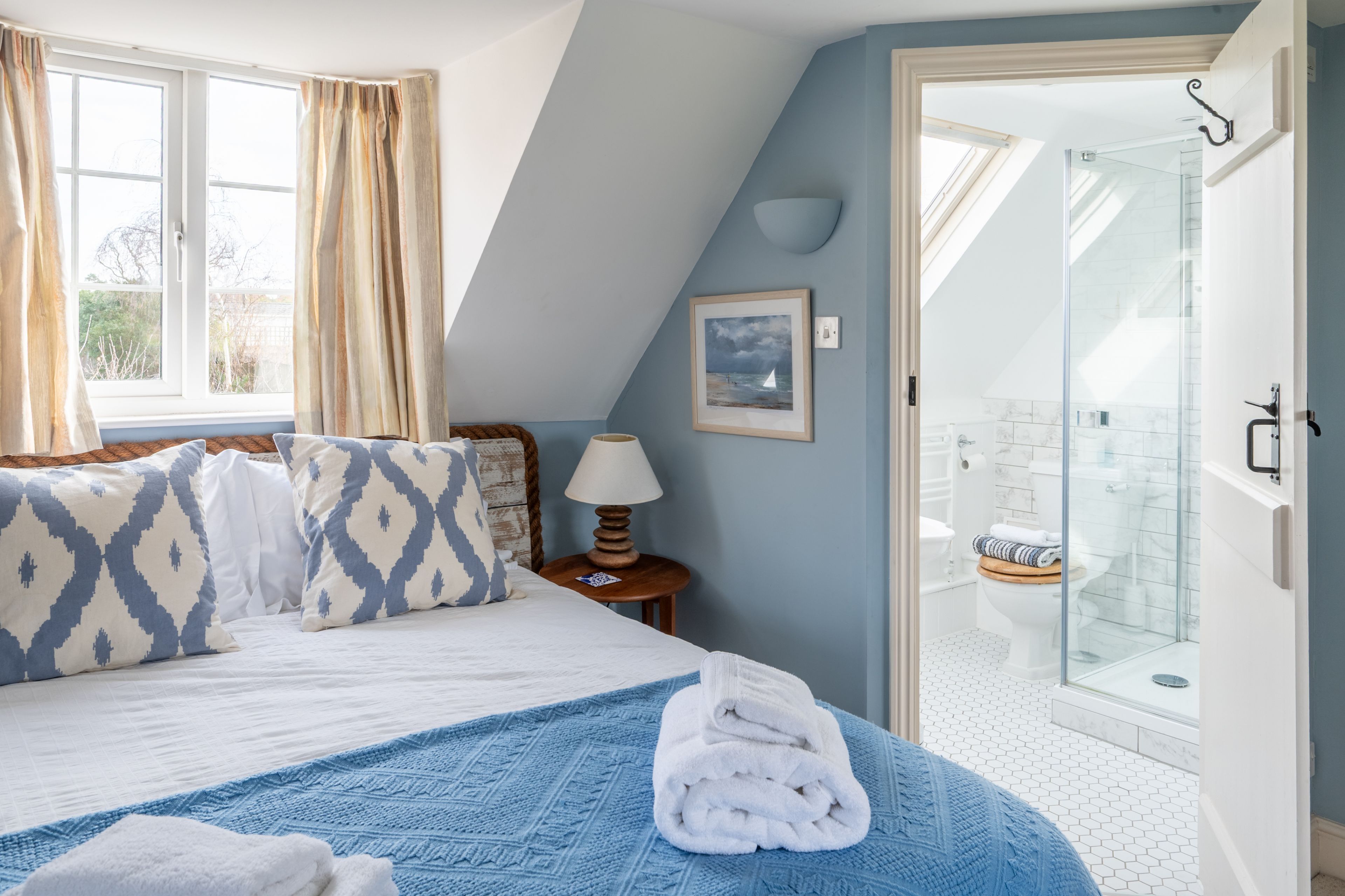 A bedroom with a bed, pillows, a bedside table with a lamp, a folded towel, and a view of a bathroom with a shower and toilet.