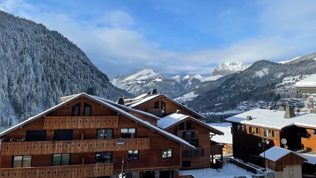Un chalet in legno con un balcone, sullo sfondo di montagne innevate e un cielo sereno.