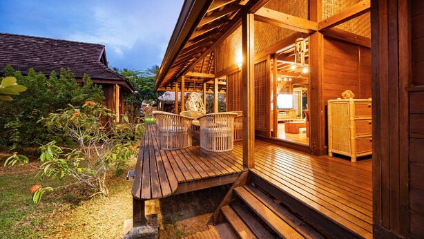 A wooden building with a porch, wicker furniture, and a thatched-roof structure in the background.