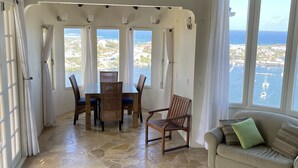 A spacious room with a stone floor, a dining area with wicker furniture, a sofa with cushions, and large windows offering a view of the ocean. - SERENDIPITY ocean views Villa Serenity & Villa Serenade A Unique Island Escape.  (Upper Prince's Quarter)