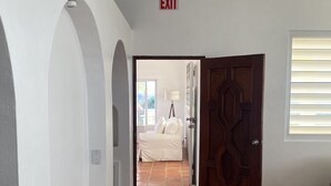Corredor com piso ladrilhado, placa de saída, porta de madeira e janela com persianas.