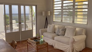 Uma sala de estar espaçosa com um sofá branco, uma mesa de centro e um abajur. Há grandes janelas com persianas, um ventilador de teto e vista para o exterior.