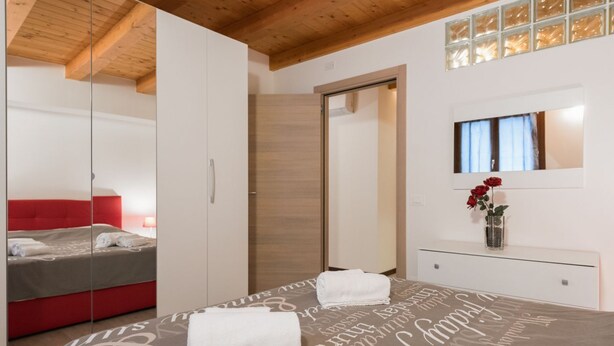 A bedroom with a bed, a red sofa, a mirror, a wooden wardrobe, and a window with a view. - Ampio alloggio con due camere da letto (Verona)