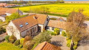 Un bâtiment en pierre, surmonté d’un toit en tuiles, entouré de champs verdoyants et d’arbres.