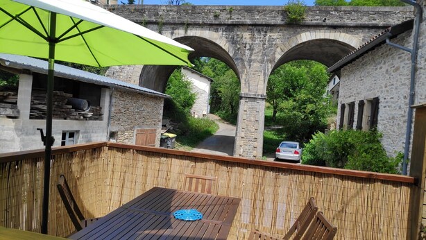 A patio area with a table and chairs, a green umbrella, and a stone bridge in the background. - Parsley Holiday Home, Tarn, South West of France (Cambounès)