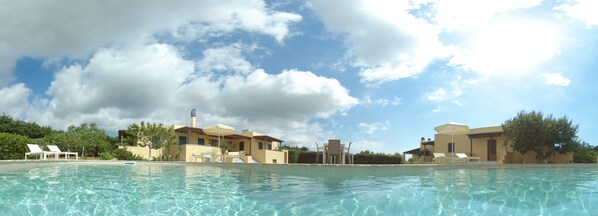 A pool area with clear water, lounge chairs, and buildings with a Mediterranean architectural style.