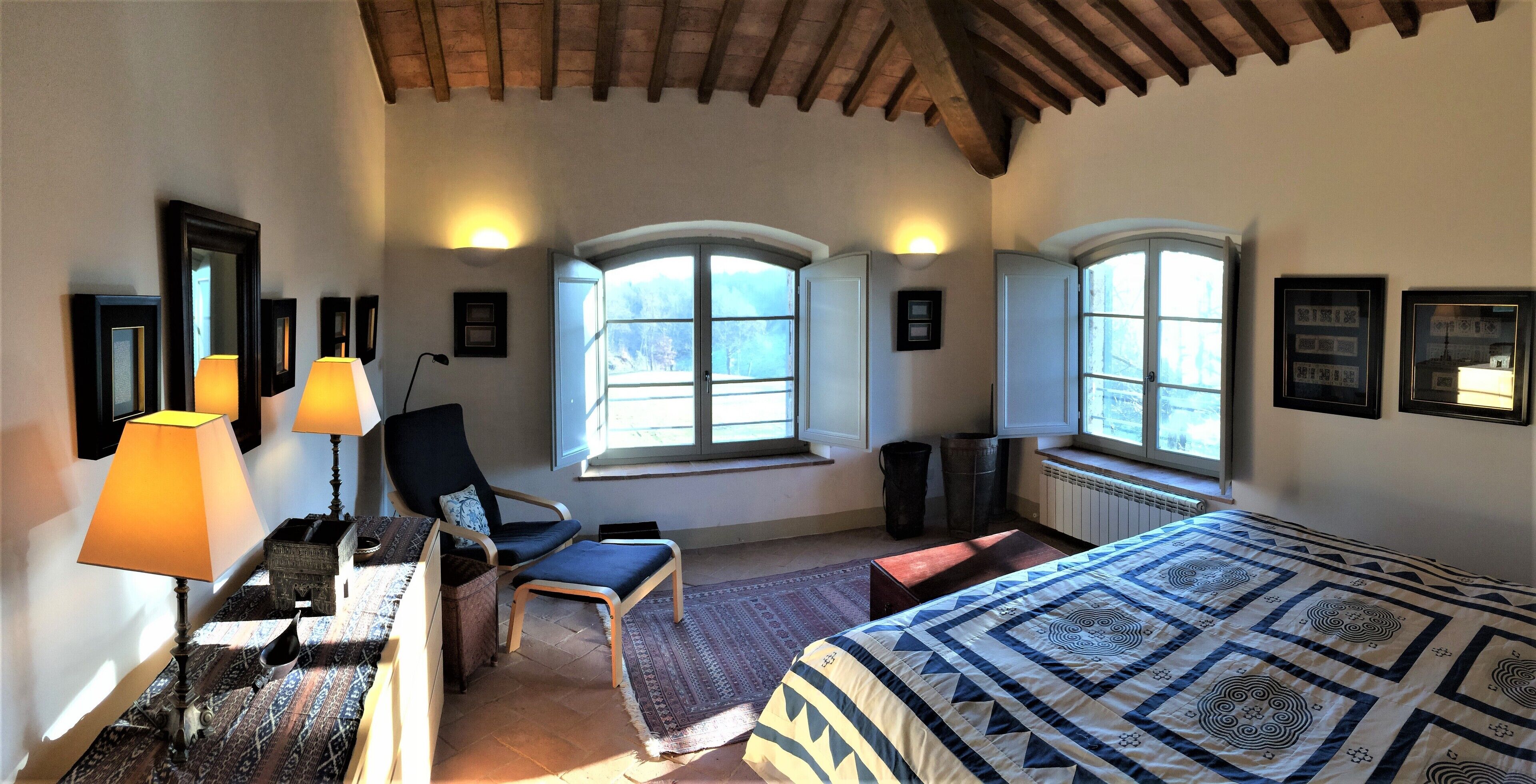 Un dormitorio con cama, una silla, un escritorio con lámpara y ventanas amplias.