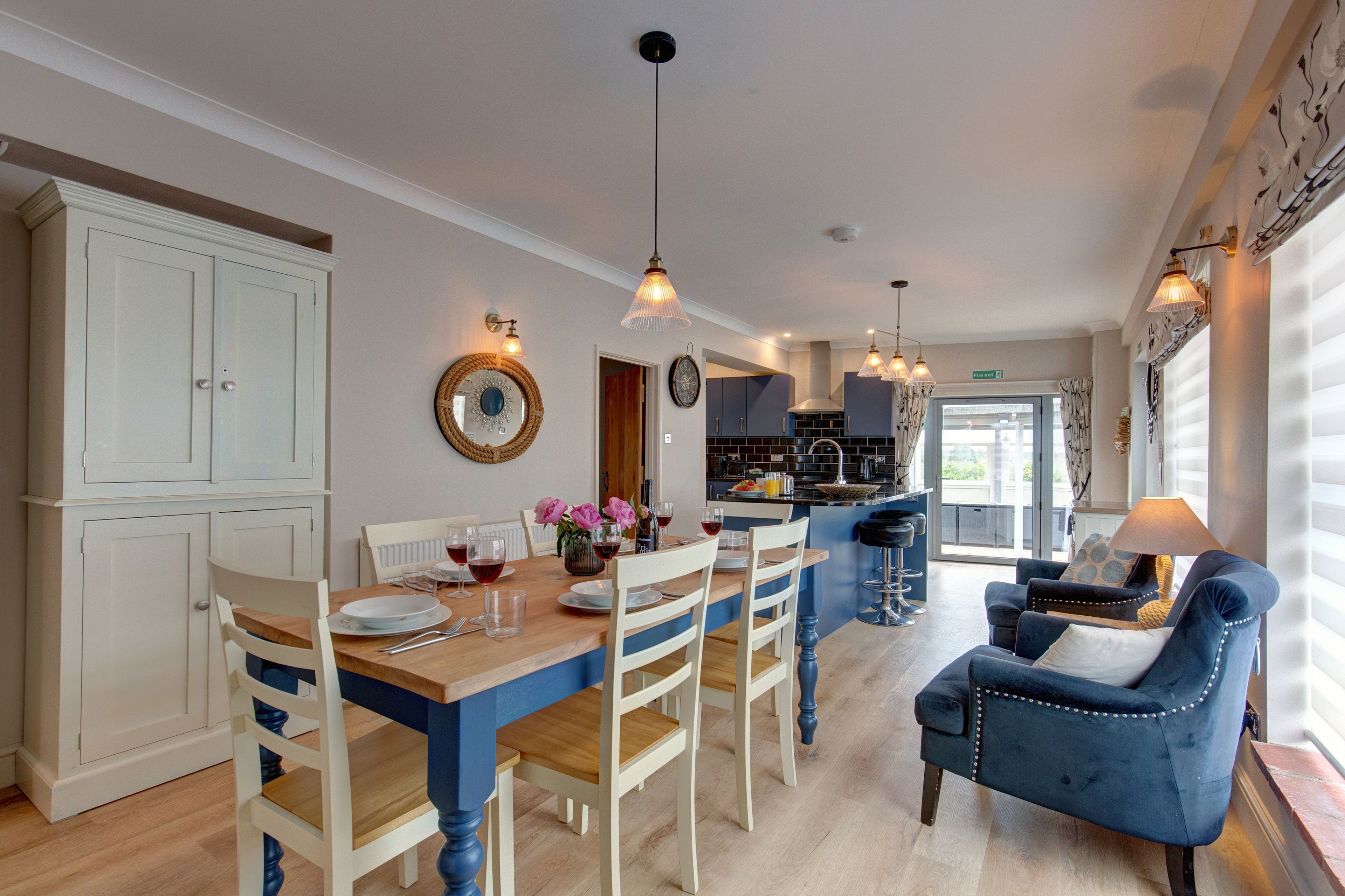 Een moderne keuken met witte kasten, een houten eettafel en blauwe stoelen. In de woonkamer staat een blauwe fauteuil en een lamp.