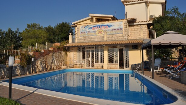 Une piscine avec une terrasse carrelée, un espace de repos couvert et un bâtiment orné d’une fresque murale.
