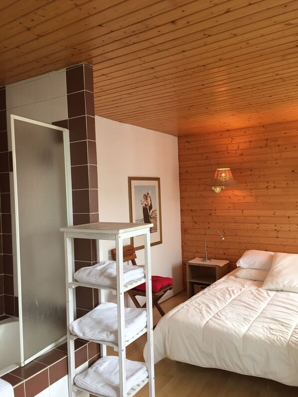 A bedroom with a wooden ceiling, a bed with white bedding, a white shelf with towels, a framed picture on the wall, and a small table with a lamp. - NEW !! Apartment in the heart of granville with panoramic view (Granville)