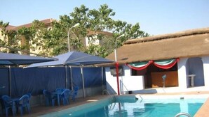 A swimming pool with blue chairs and umbrellas, a thatched roof structure, and a clear sky. - Nugget Hotel (Abuja)