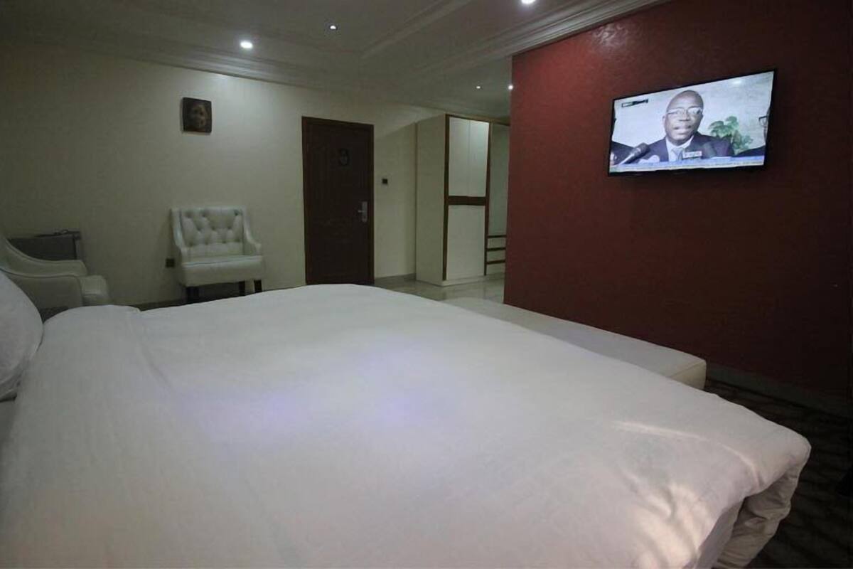 A hotel room with a large bed, a white chair, a television on the wall, and a door leading to another room.