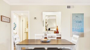 Een eethoek met een houten tafel, witte stoelen en een schaal eten. Er hangen decoraties aan de muur en erboven bevindt zich een spiegel.