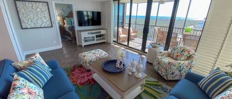 Un salón amplio con televisor grande, ventilador de techo y balcón con mobiliario exterior.