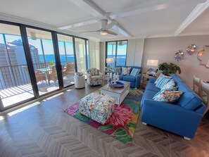 Un salon spacieux doté d’une grande porte-fenêtre coulissante donnant sur un balcon, d’un ventilateur de plafond, d’un canapé bleu et d’un tapis aux couleurs vives.