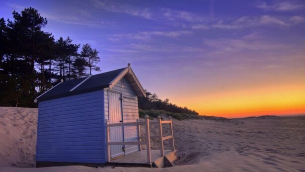 A small beach hut with a sloped roof and a wooden door, situated on a sandy beach with a clear sky in the background. - Angel Cottage, Great Walsingham, Norfolk (Walsingham)