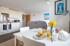 Une cuisine moderne avec des armoires blanches, un four à micro-ondes et un four traditionnel. On y trouve une table à manger avec des aliments pour le petit-déjeuner, un vase rempli de fleurs jaunes et un tableau encadré accroché au mur.