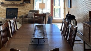 Un comedor con una gran mesa de madera, sillas, una lámpara de araña, una chimenea y un televisor montado en la pared.