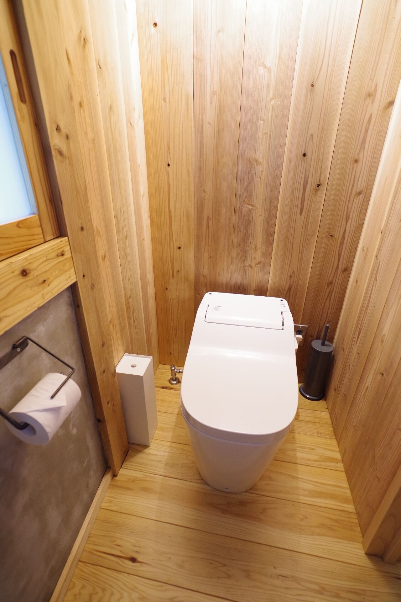 A wooden-paneled bathroom with a toilet, a toilet paper holder, and a trash can.
