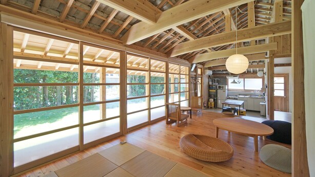 Una habitación amplia con ventanas grandes, vigas de madera, esteras de tatami y una zona de cocina.
