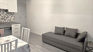 A modern, minimalist living space with a grey sofa, a dining table with chairs, and a kitchen area with white cabinets and a tiled backsplash. - Appartamento con due camere a 1 km da Piazza Erbe (Verona)