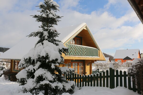 A wooden cabin with a green roof, surrounded by snow-covered trees and a picket fence.