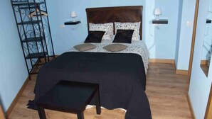 Un lit bien fait, recouvert d’une couvre-lit sombre, avec deux oreillers et une tête de lit en bois. Une petite table se trouve devant le lit, et une étagère en métal est placée à gauche.