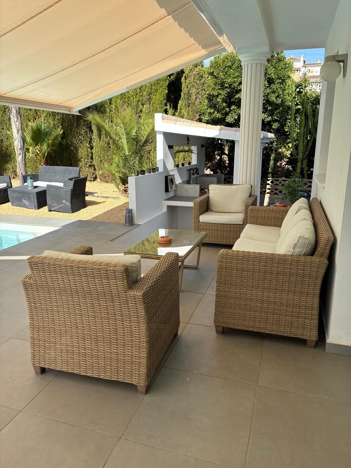 A covered outdoor seating area with wicker furniture, a pool, and a view of greenery.