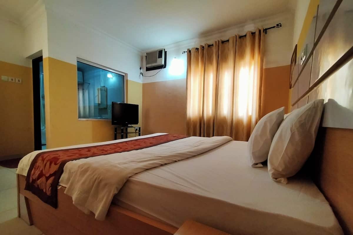 A hotel room with a bed, a television, a window with curtains, and an air conditioning unit.