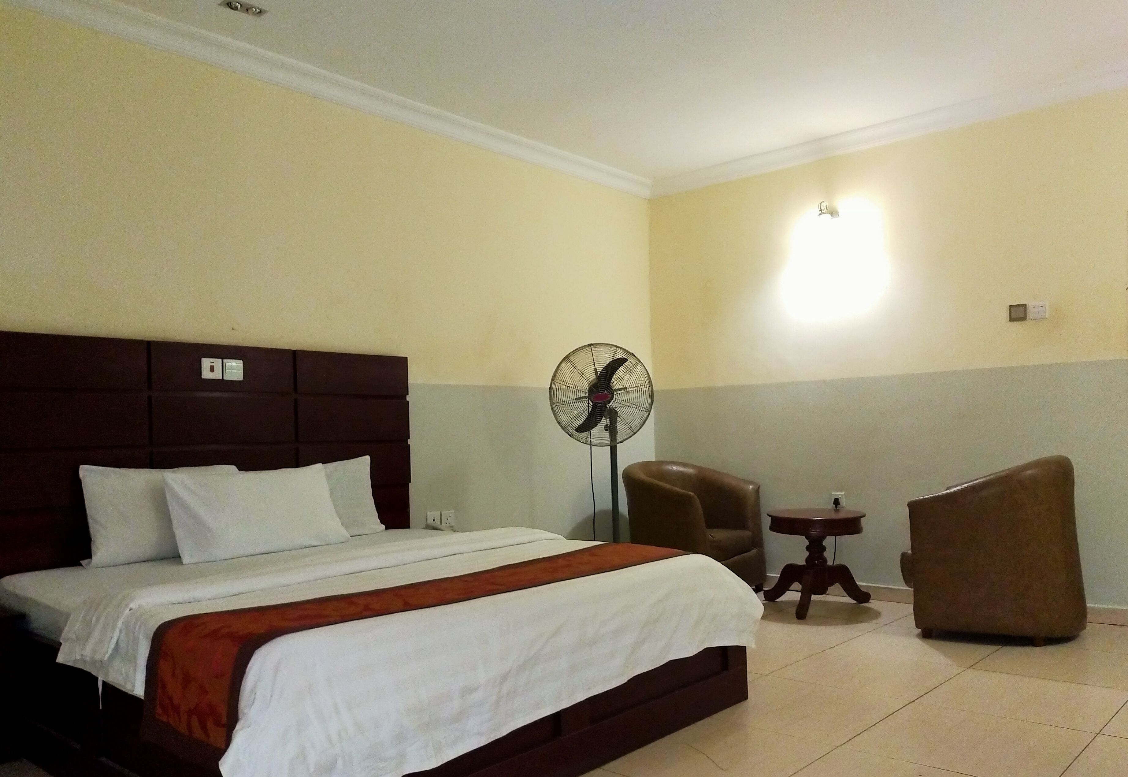 A hotel room with a large bed, a standing fan, two armchairs, and a small table.