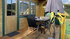 Une terrasse en bois avec une table dressée pour les repas, des chaises d’extérieur et un grand parasol.
