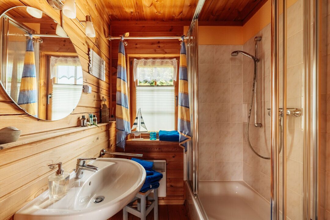 A wooden cabin bathroom with a sink, mirror, shower, and a window with curtains.