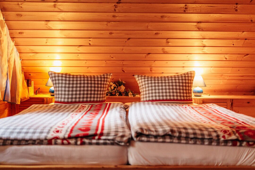 Une pièce chaleureuse aux murs en bois, avec deux lits dotés de draps à carreaux, une table de chevet avec une lampe et une petite table ornée de fleurs.
