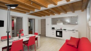 Cocina moderna con armarios blancos, microondas y estufa. Hay un comedor con mesa y sillas rojas. El techo tiene vigas de madera a la vista.