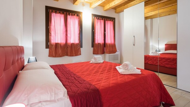 Un dormitorio con una colcha roja, dos ventanas con cortinas marrones, un armario blanco y un espejo.