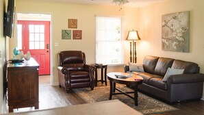 A living room with a brown leather chair, a brown leather sofa, a small round table, and a lamp. There is a red door, a window with blinds, and a ceiling fan.