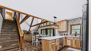 A spacious kitchen with wooden cabinets, a large island, and a dining area with white chairs. - Freshly Remodeled Cabin Escape—Hot Tub, Treehouse, Game Room, WiFi, 2 Decks (Rockbridge)