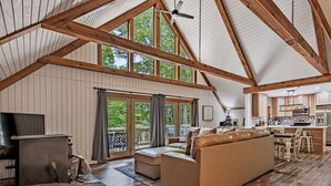 A spacious living area with a high ceiling, wooden beams, a large sofa, a television, and a kitchen area. - Freshly Remodeled Cabin Escape—Hot Tub, Treehouse, Game Room, WiFi, 2 Decks (Rockbridge)
