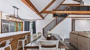 A modern kitchen with a central island, wooden beams, a staircase, and a dining area with a glass table and white chairs. - Freshly Remodeled Cabin Escape—Hot Tub, Treehouse, Game Room, WiFi, 2 Decks (Rockbridge)