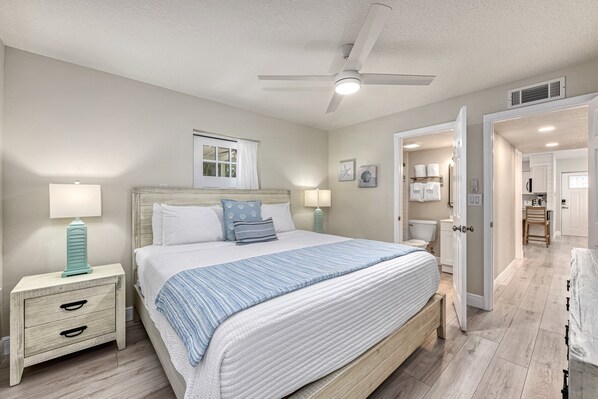 Une chambre à coucher avec un lit, des tables de chevet, un ventilateur de plafond et une salle de bain visible à travers une porte ouverte.