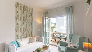 Una sala de estar moderna con sofá, sillón y mesa de centro, un balcón con vistas a edificios y montañas, y un ventanal con cortinas.