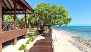 Une terrasse en bois qui mène à une plage, avec un grand arbre et un bâtiment doté d’un espace de repos abrité.