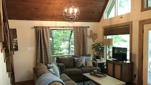 Una sala de estar acogedora con techo de madera, un sofá, una mesa de centro, un televisor y un ventanal con cortinas.