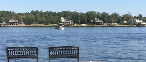 A waterfront view with two benches on a dock, calm water, and houses in the background.