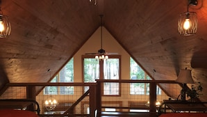 Un acogedor dormitorio en el ático con vigas de madera, techo inclinado y un amplio ventanal con vistas a árboles. Hay dos camas con ropa de cama roja, una mesita y una lámpara.