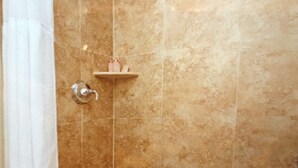 Eine Dusche mit beigen Fliesen, einem weißen Vorhang und einer kleinen Ablage für Toilettenartikel.