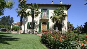 Um edifício de dois andares com varanda, rodeado por palmeiras e um jardim bem cuidado com flores vermelhas.