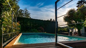 Piscina retangular com um deck de madeira, rodeada por vegetação exuberante e um céu claro.